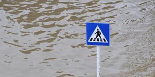 Ein Verkehrszeichen "Fussgängerübergang" ragt auf einer überschwemmten Strasse aus den Fluten hervor. (Foto: picture-alliance / Romain Fellens | Romain Fellens)