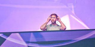 Calvin Harris steht auf der Bühne am DJ-Pult und setzt sich Kopfhörer auf. (Foto: picture alliance / Photoshot | -)