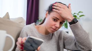 ine junge Frau sitzt in einem Wohnzimmer und schaut besorgt auf ihr klingelndes Smartphone (Foto: dpa)