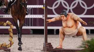 Ein Pferd springt gerade über ein Hindernis. Daneben steht eine Sumo-Statue (Foto: picture alliance/dpa | Friso Gentsch)