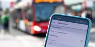 Auf einem Handy ist die Buchungsseite des 9-Euro-Tickets aufgerufen mit einem roten Bus im Hintergrund. (Foto: picture alliance/dpa | Melissa Erichsen)