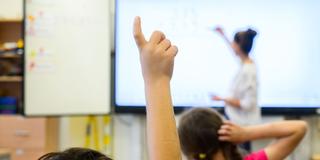 Ein Schüler meldet sich per Handzeichen während eine Lehrerin auf eine digitale Schultafel im Klassenraum einer 4. Klasse einer Grundschule schreibt (Foto: picture alliance/dpa | Julian Stratenschulte)