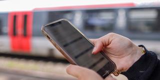 Frau mit iPhone in der Hand. Im Hintergrund eine Bahn. (Foto: picture alliance/dpa | Jan Woitas)