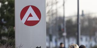 Stele mit dem Zeichen, Logo der Agentur für Arbeit vor einem Gebäude. Personen gehen daran vorbei.  (Foto: picture alliance / imageBROKER | Jan Tepass)