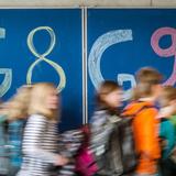 Eine Tafel, auf der G8 und G9 steht mit Schülern die vor der Tafel laufen. (Foto: dpa)