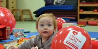 Ein Kleinkind spielt auf dem Boden vor einem Herzenssache-Luftballon (Foto: Herzenssache e.V.)