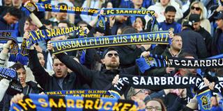 Fans des 1. FC Saarbrücken stehen auf der Tribüne und halten ihre Schals hoch und singen gemeinsam die Vereinshymne im Ludwigsparkstadion (Foto: IMAGO / Fussball-News Saarland)