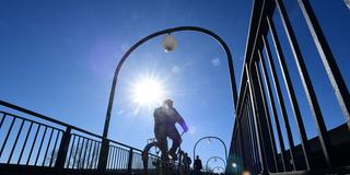 Fußgänger und ein Radfahrer überqueren bei warmem Wetter eine Brücke. (Foto: IMAGO / BeckerBredel)