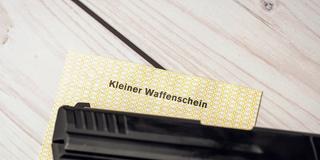 Ein kleiner Waffenschein liegt auf einem Tisch unter dem Lauf einer Pistole (Foto: IMAGO / Bihlmayerfotografie)