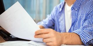 Jemand sitzt am Schreibtisch und hält ein Blatt Papier in der Hand (Foto: IMAGO / Zoonar)