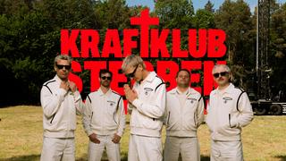 5 Männer in weißen Anzuügen stehen auf einer trockenen Wiese. Im Hintergrund ist ein roter "Kraftklub"-Schriftzug zu sehen. (Foto: Philipp Gladsome)