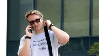 Naarly läuft mit hellem T-Shirt, Sonnenbrille, und Handtasche vor einem Gebäude vorbei und telefoniert. (Foto: @naarl_y (Instagram))