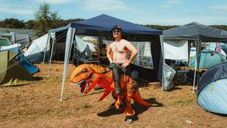 Ein Festivalbesucher steht oberkörperfrei auf dem Campingplatz, er trägt ein Kostüm, das aussieht, als würde er auf einem Dino reiten. (Foto: MxM Photo)