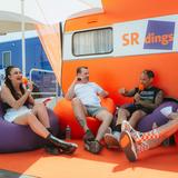 Vier Menschen sitzen auf Sitzsäcken vor einem orangenen SRdings-Wohnwagen (Foto: MxM Photo)