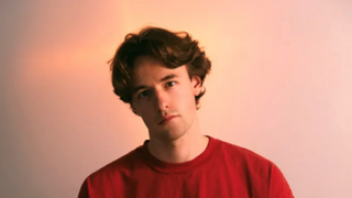 Ein junger Mann mit mittellangen braunen Haaren steht vor einem hellen Hintergrund. Er trägt ein rotes T-Shirt und guckt mit neutralem Gesichtsausdruck in die Kamera. (Foto: Pressefoto/VIOSIONV)